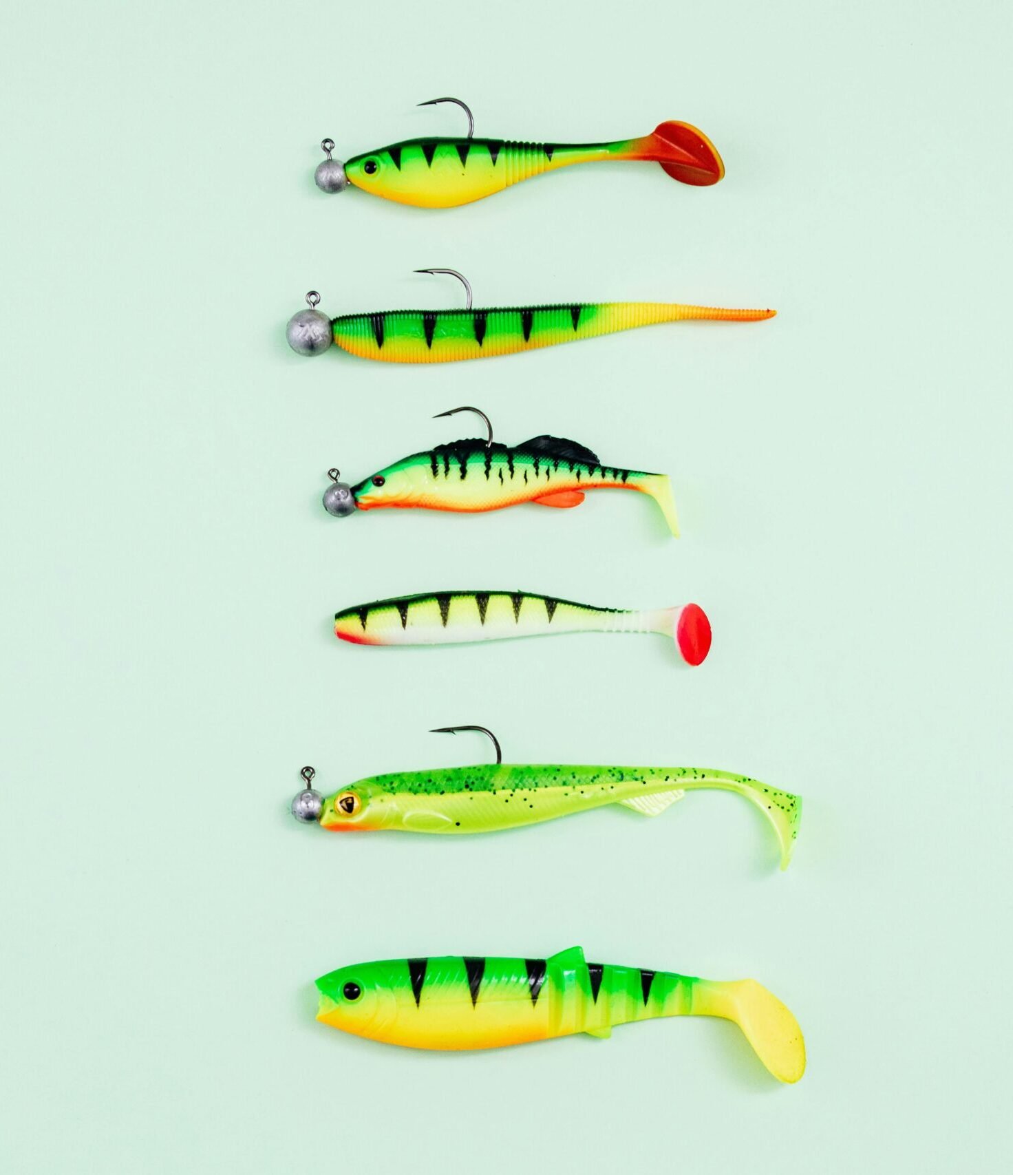 Six brightly colored soft plastic fishing lures with jig heads arranged vertically on a light green background.